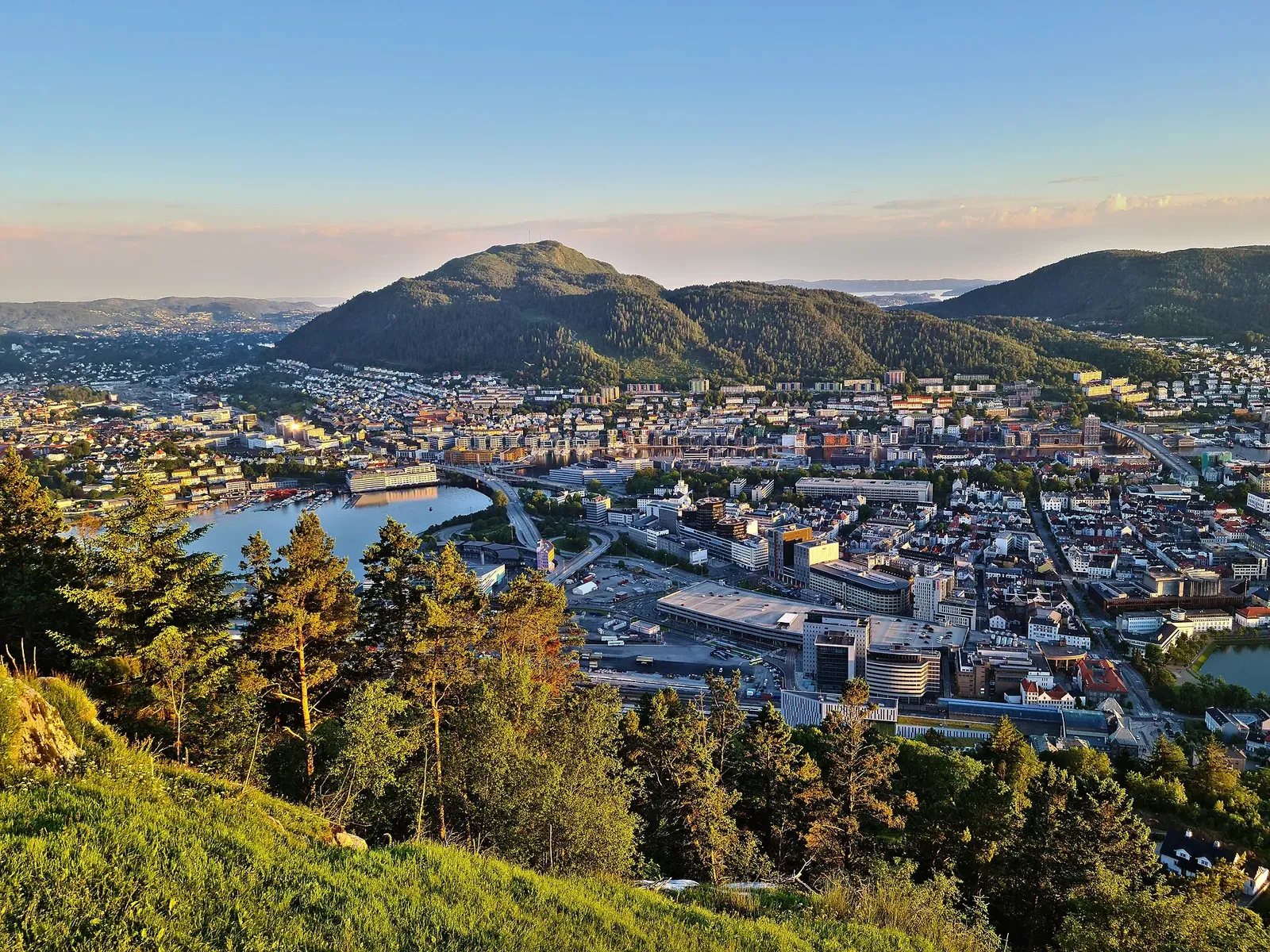 Vue plongeante sur une ville au pied de collines boisées, avec un bras d'eau, à la lumière dorée du soir