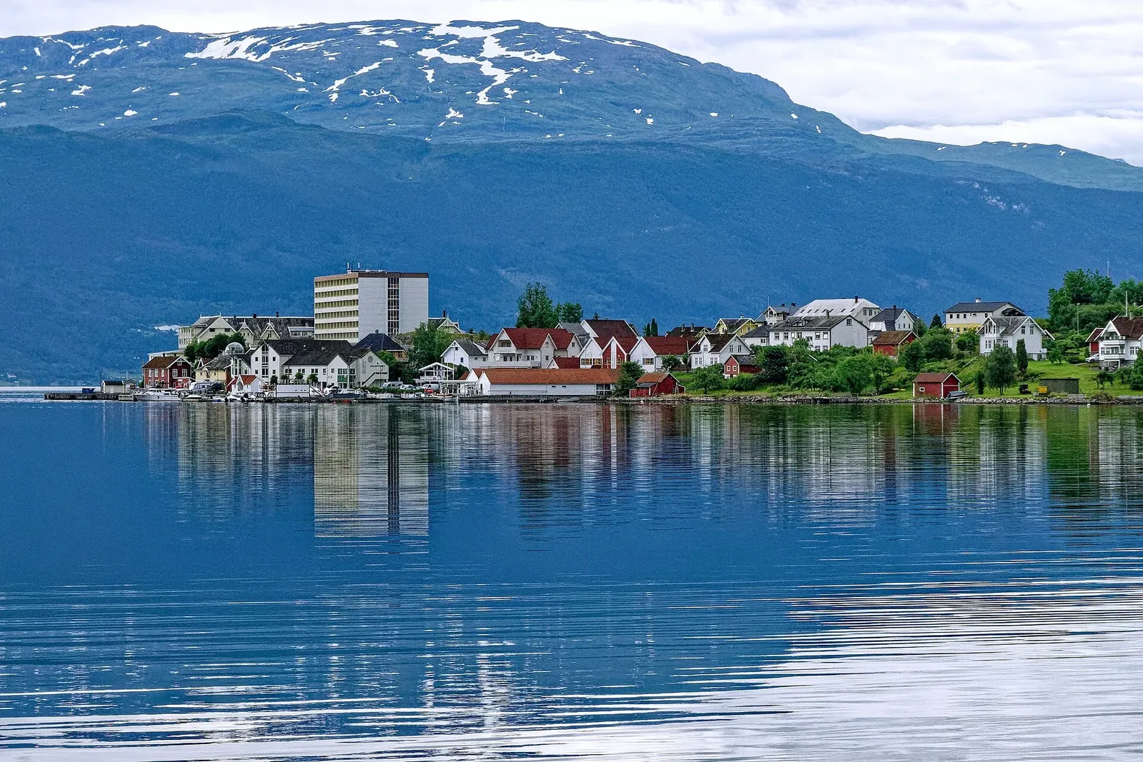 Village de maisons blanches et rouges au bord d'un fjord calme, avec des montagnes aux névés en arrière-plan