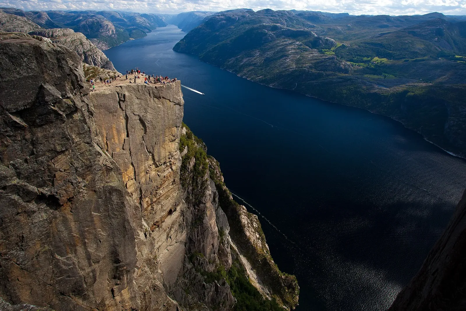 Vue plongeante sur un fjord bleu sombre bordé de falaises, avec un plateau rocheux où se tiennent des visiteurs