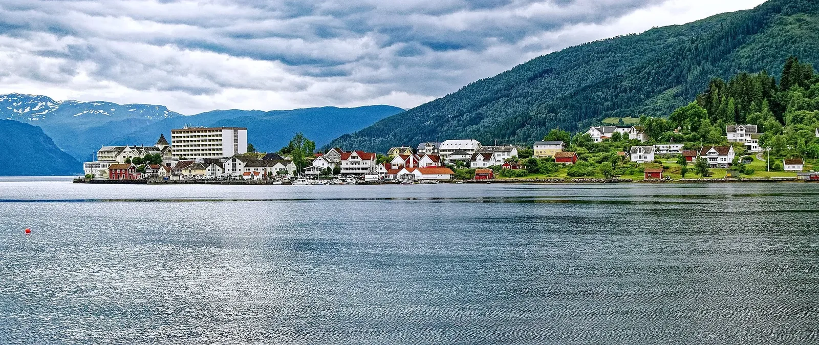 Village aux maisons blanches et rouges au bord d'un fjord calme, entouré de montagnes boisées et de sommets enneigés au loin