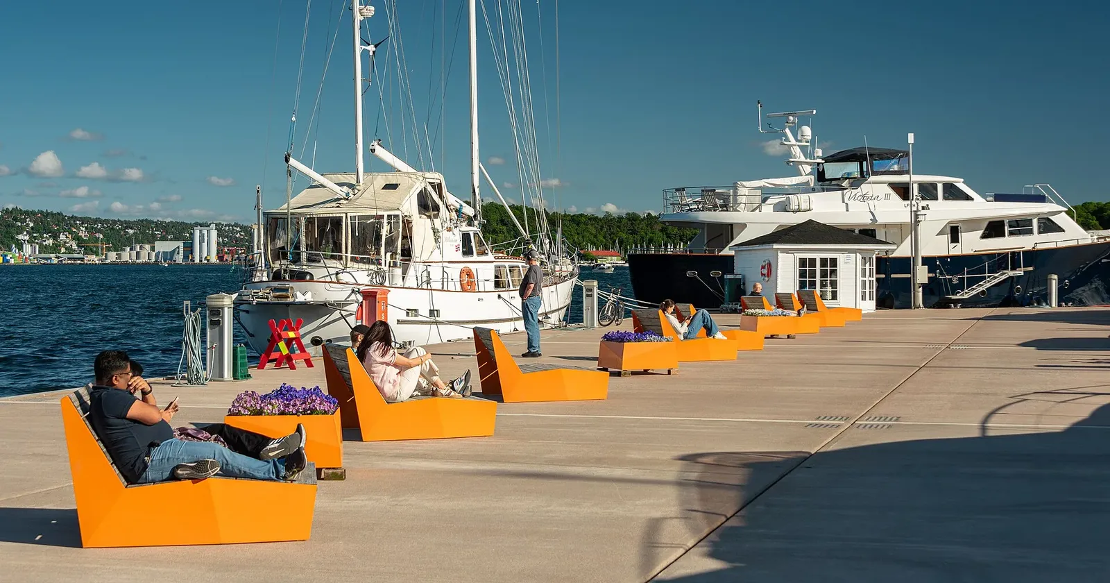 Quai ensoleillé d'une marina avec voiliers et yacht amarrés, bancs orange où des gens se détendent, fjord en arrière-plan