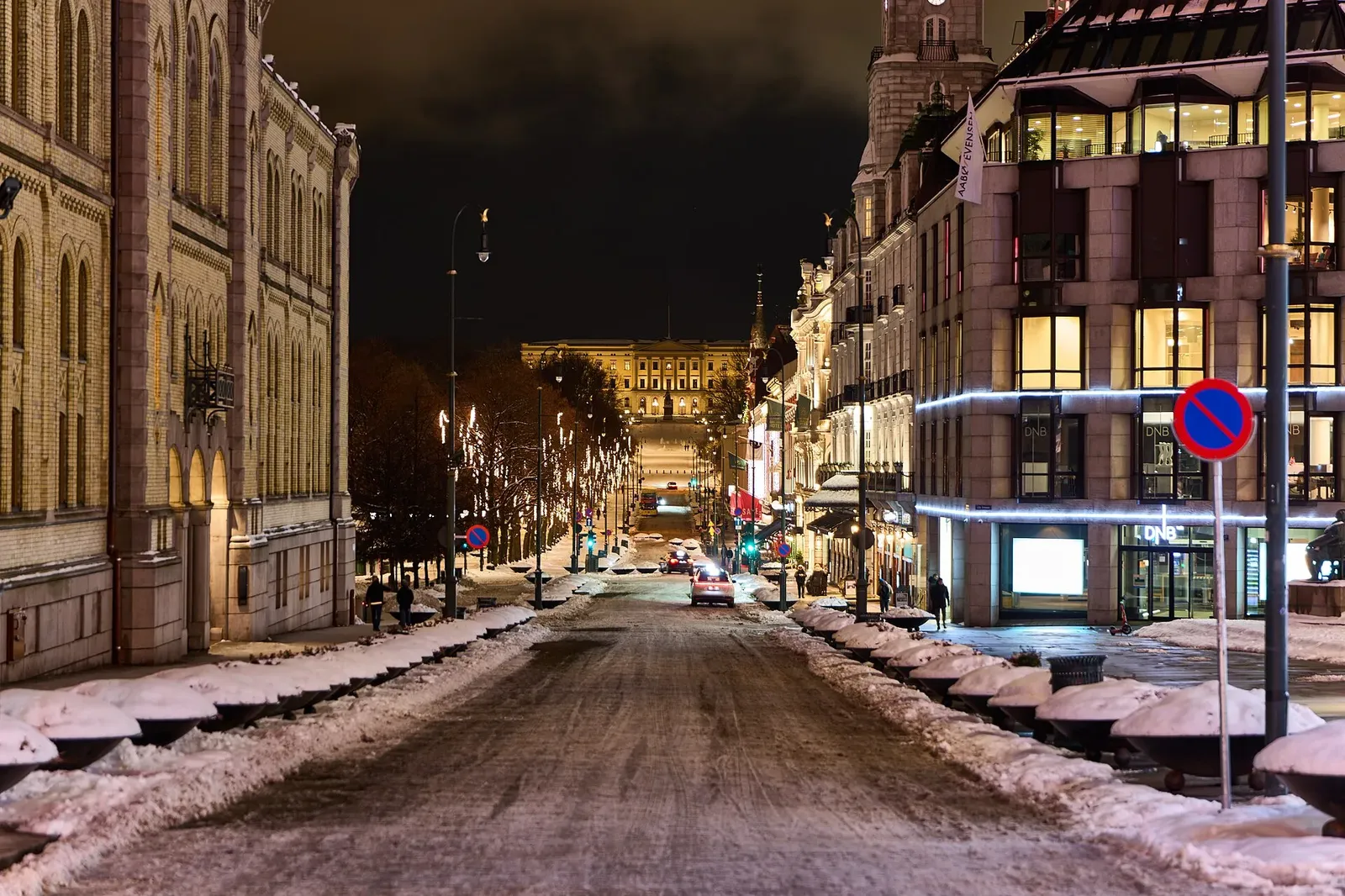 Rue enneigée de nuit, bordée de façades éclairées, avec un grand bâtiment illuminé au bout de la perspective