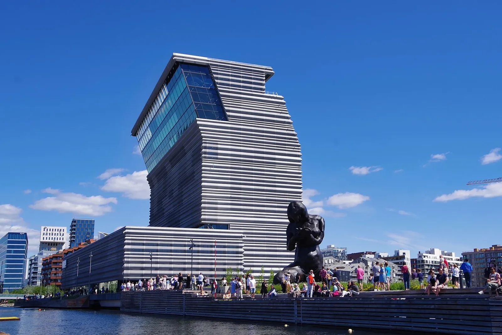 Façade inclinée du musée Munch au bord de l'eau, avec une grande sculpture sombre et des visiteurs sous un ciel bleu