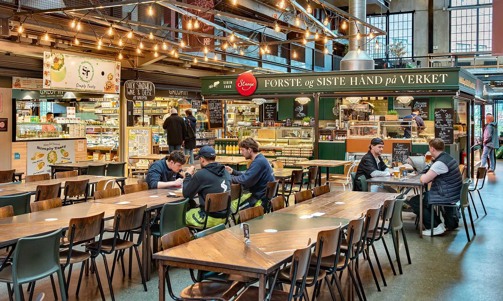 Intérieur d'une halle alimentaire avec stands de restauration, longues tables en bois et clients attablés sous des guirlandes lumineuses