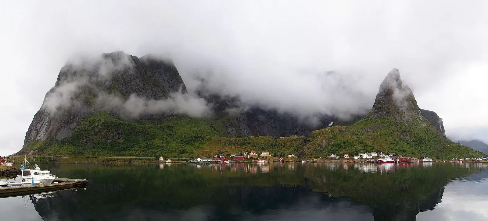 Panorama de montagnes aux sommets embrumés qui se reflètent dans l'eau calme d'un fjord, avec des maisons au pied et un bateau à quai