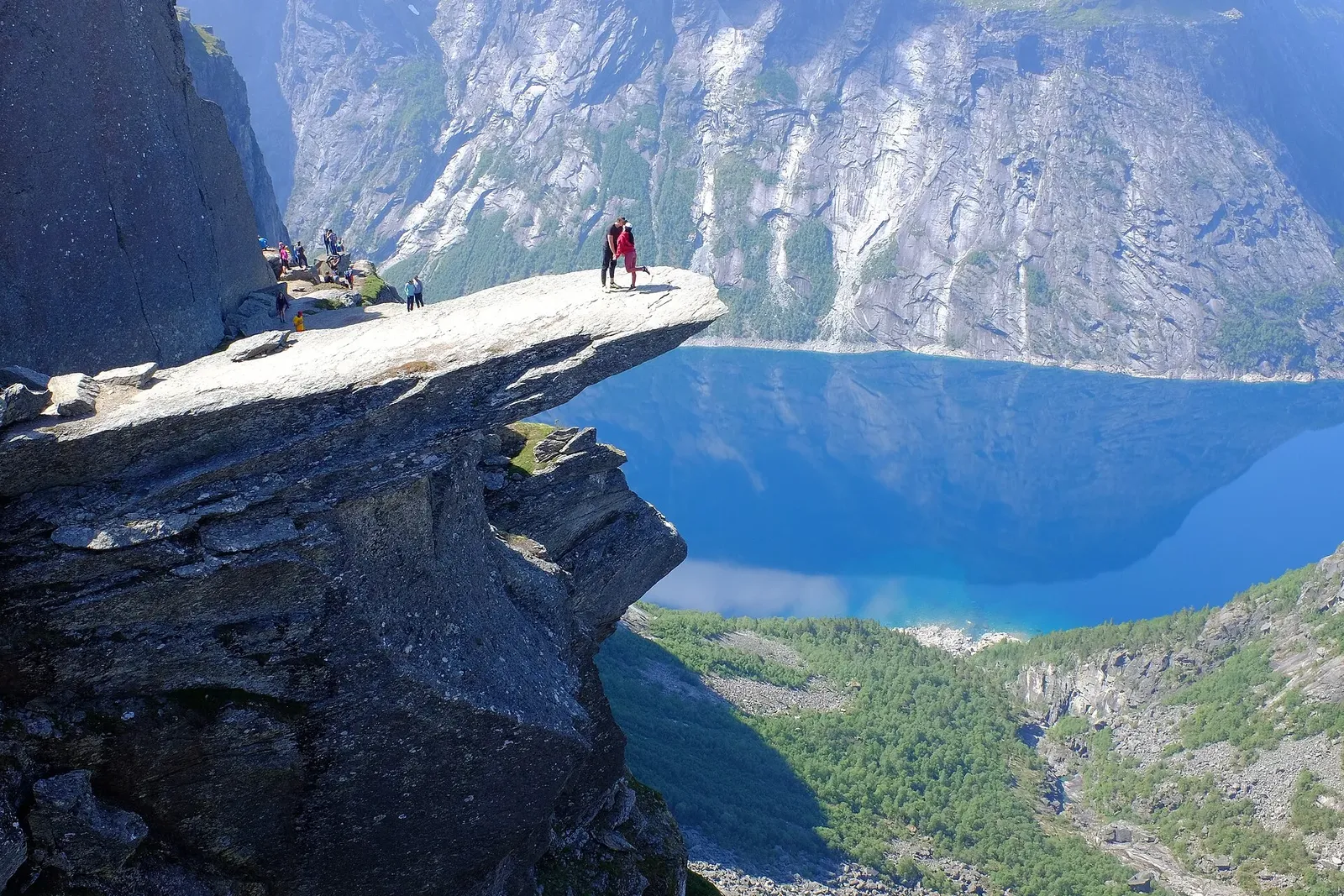 Rocher en surplomb au bord d'une falaise, avec des randonneurs, au-dessus d'un lac bleu encaissé entre des parois rocheuses